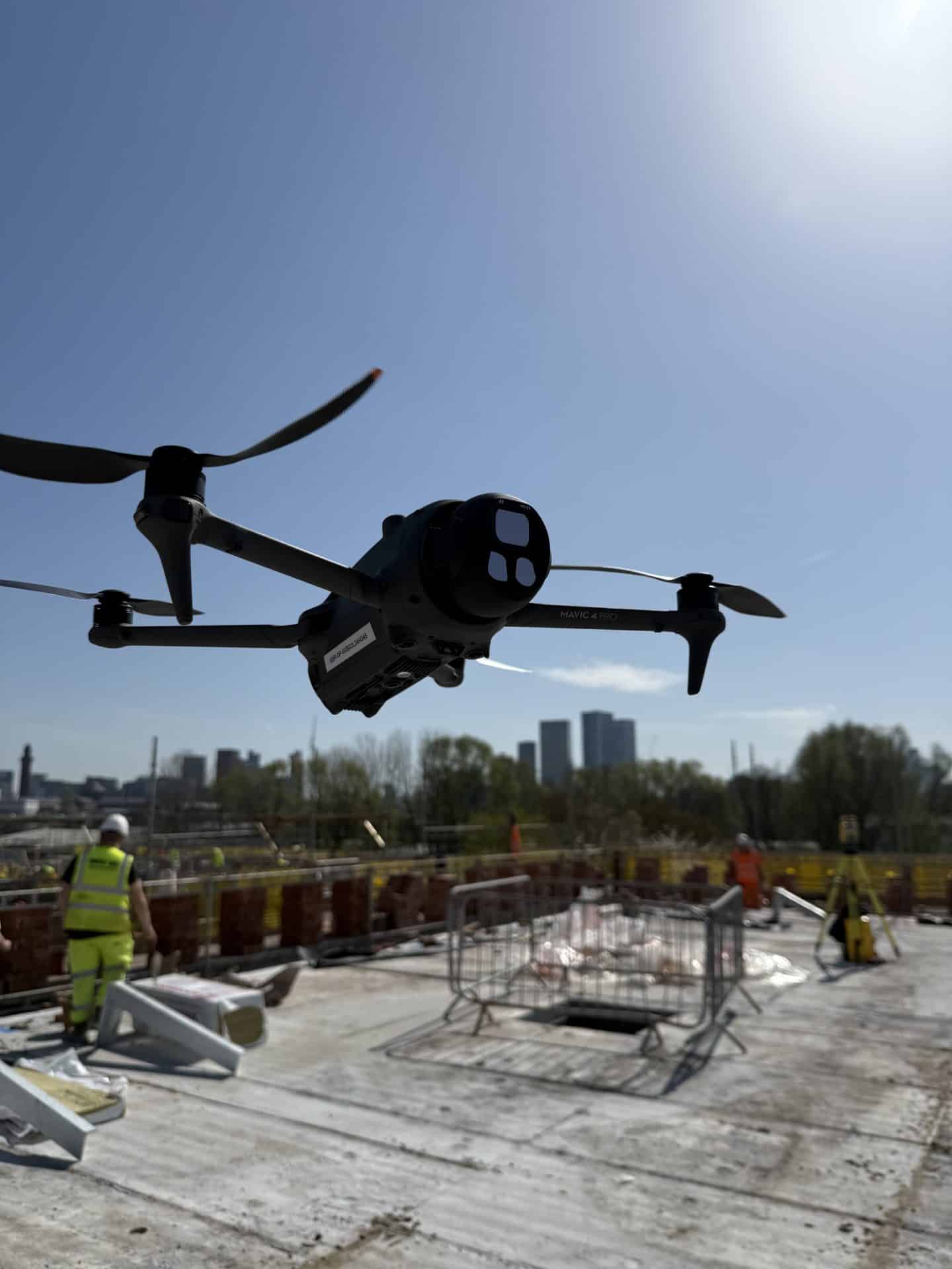 Drone flying over construction site with workers and city skyline in background.